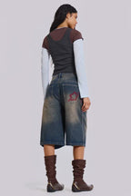 Back view of a woman wearing dark wash wide leg cropped denim culottes with red embroidered detail on the back pocket, a dark grey tank top layered over a light blue long-sleeve shirt, and brown boots.