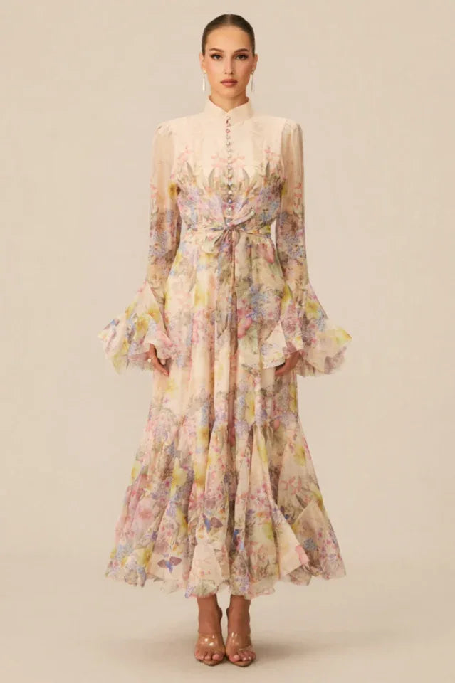 Woman wearing a long, flowing floral print chiffon dress with a high neckline, long bell sleeves, and a tiered ruffled skirt.