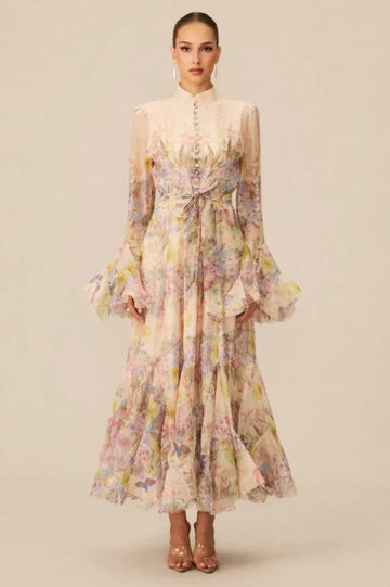 Woman wearing a long, flowing floral print chiffon dress with a high neckline, long bell sleeves, and a tiered ruffled skirt.