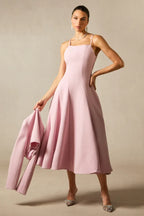 Woman holding a pink jacket while wearing a pink formal dress with thin straps and a flared skirt.