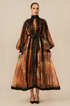 Woman wearing a long, flowing A-line dress with a high neckline, puffed sleeves, and a black ribbon belt.