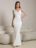 Woman in a white satin mermaid gown with a cowl neckline and a flowing one-shoulder detail, standing in front of a sofa.