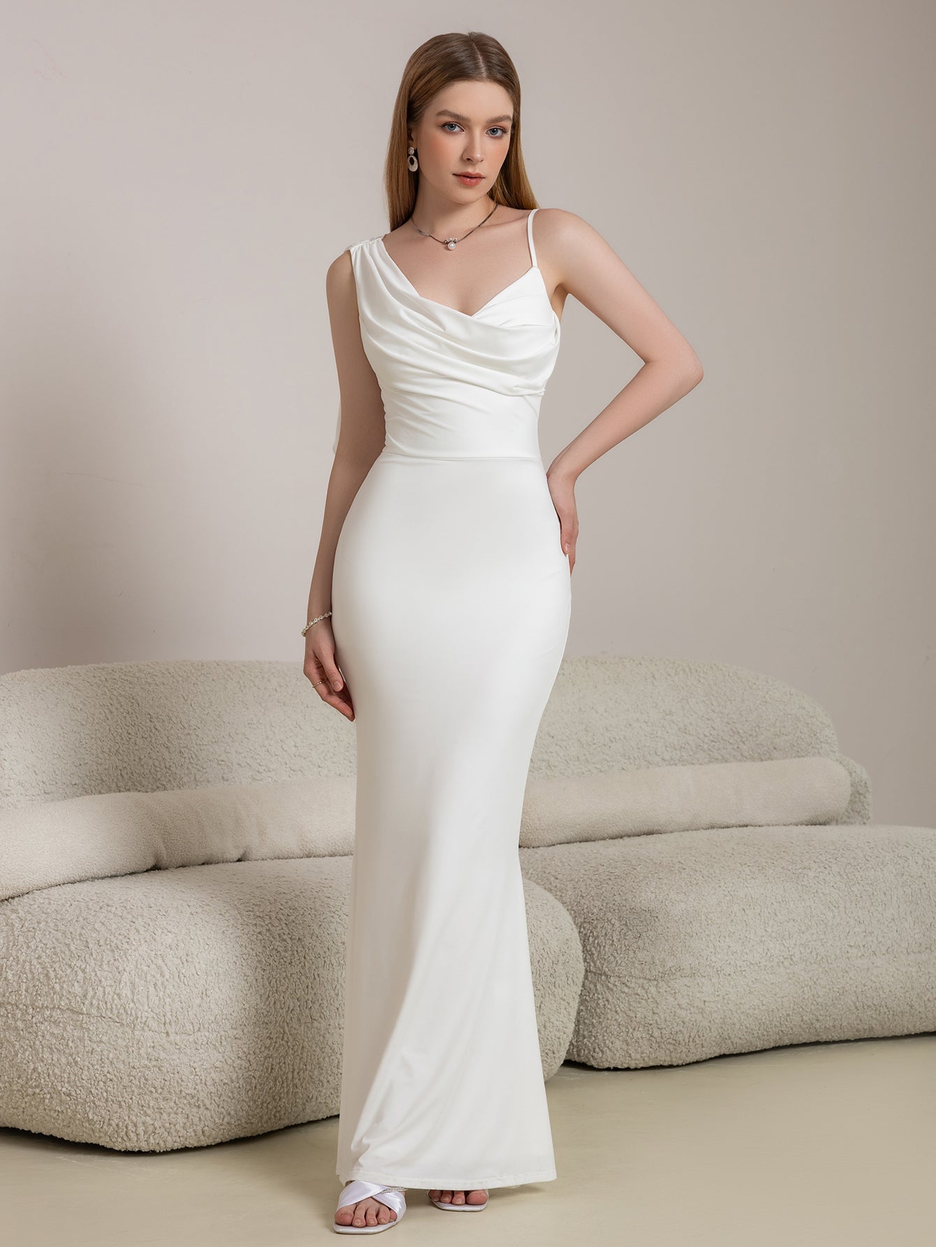 Woman in a white satin mermaid gown with a cowl neckline and a flowing one-shoulder detail, standing in front of a sofa.