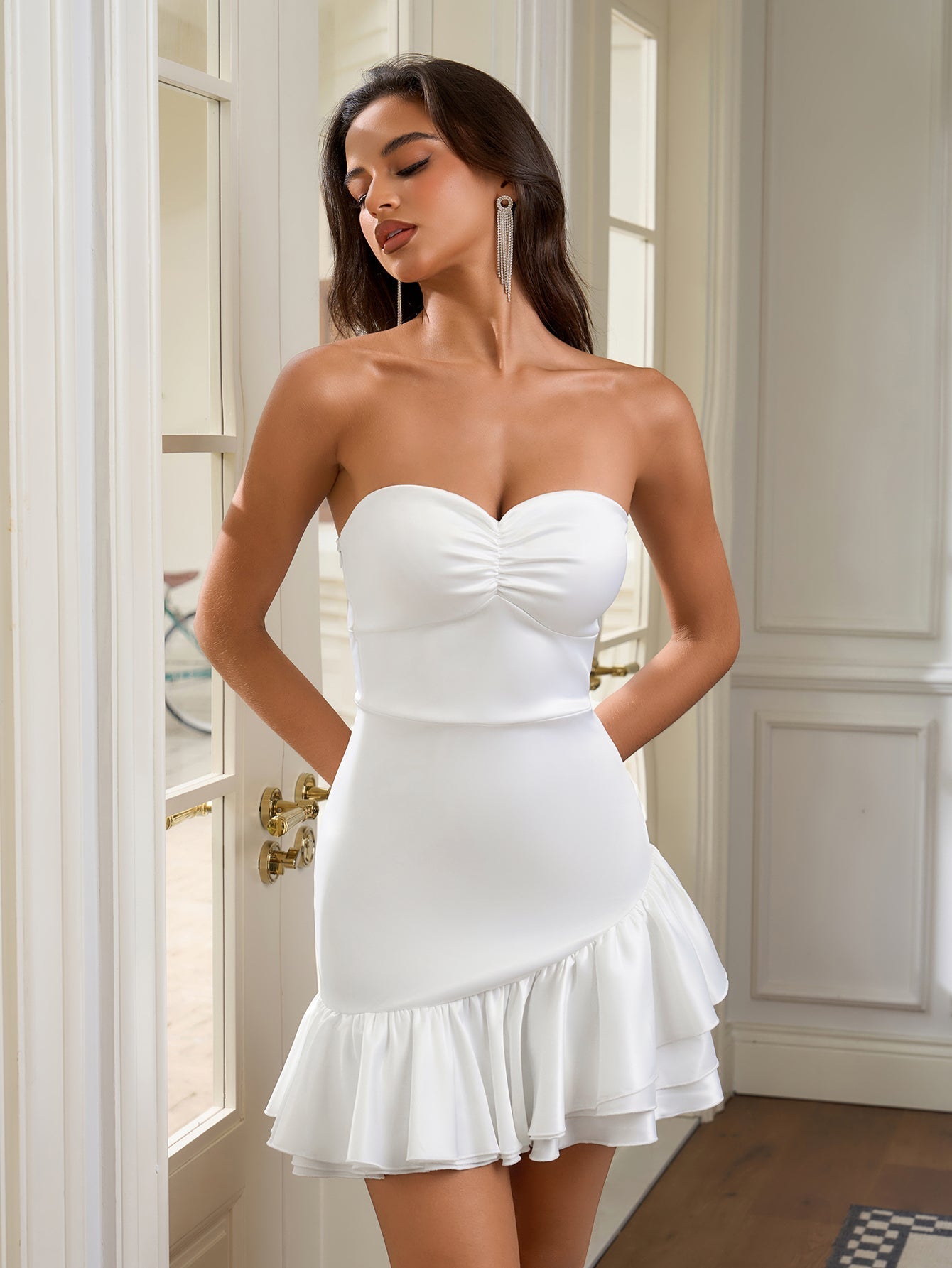 Front view of a woman in a white strapless ruched bodice sheath dress with an asymmetrical ruffle hem.