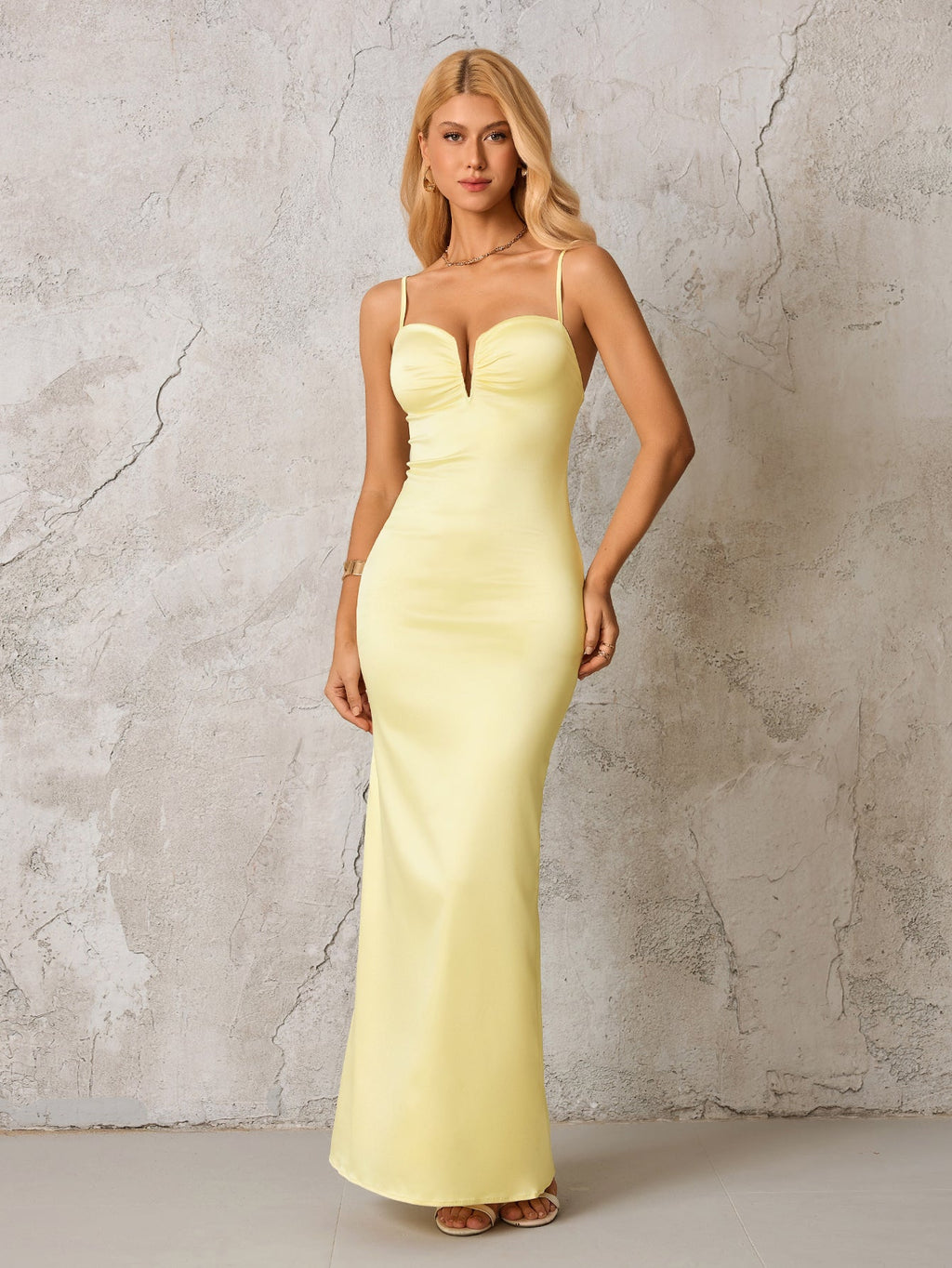 Woman in a pale yellow satin formal gown with spaghetti straps and a plunging neckline, standing against a textured gray wall.