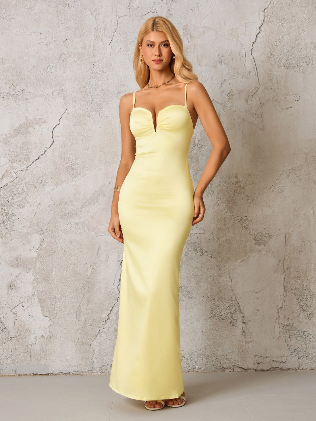 Woman in a pale yellow satin formal gown with spaghetti straps and a plunging neckline, standing against a textured gray wall.