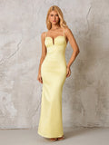 Woman in a pale yellow satin formal gown with spaghetti straps and a plunging neckline, standing against a textured gray wall.