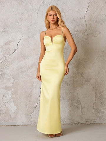 Woman in a pale yellow satin formal gown with spaghetti straps and a plunging neckline, standing against a textured gray wall.