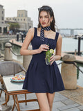Woman in a navy blue ruched bodice fit and flare dress with a square neckline, holding a drink.