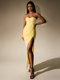Model posing in a yellow strapless evening gown with a ruched front and a dramatic slit.