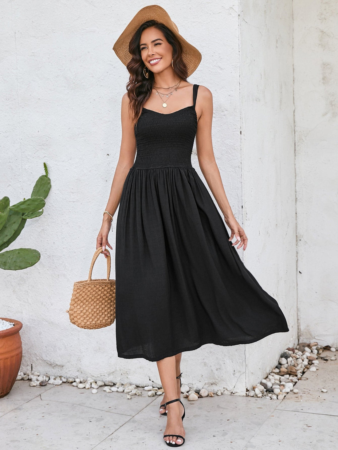 Woman wearing a black sleeveless midi dress with a smocked bodice and full skirt, holding a straw bag and wearing a straw hat.