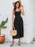 Woman in a black sleeveless midi dress with a smocked bodice and full skirt, holding a straw bag and wearing a straw hat, posing outdoors.