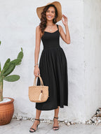 Woman in a black sleeveless midi dress with a smocked bodice and full skirt, holding a straw bag and wearing a straw hat, posing outdoors.