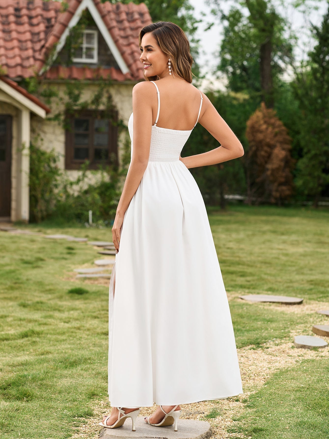 Rear view of a woman in a white maxi dress with spaghetti straps and a smocked back, standing in a garden.