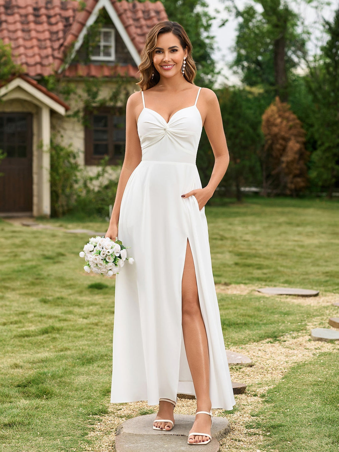 Woman wearing a white spaghetti strap maxi dress with a twisted bodice and high leg slit, holding a bouquet of white flowers.