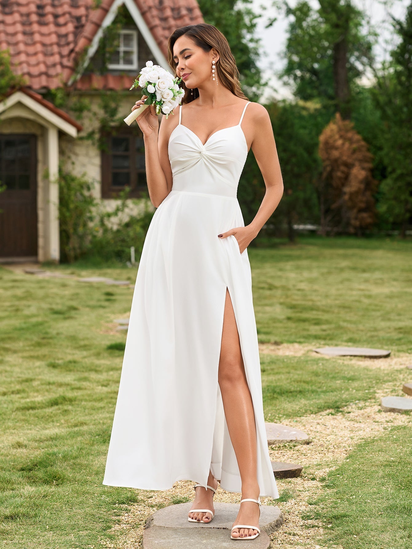 A woman in a white maxi dress with a twisted bodice and leg slit, smelling a bouquet of white flowers.