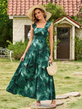 Woman wearing a green and black floral print maxi dress with ruffled shoulder straps and a square neckline, holding a straw handbag.