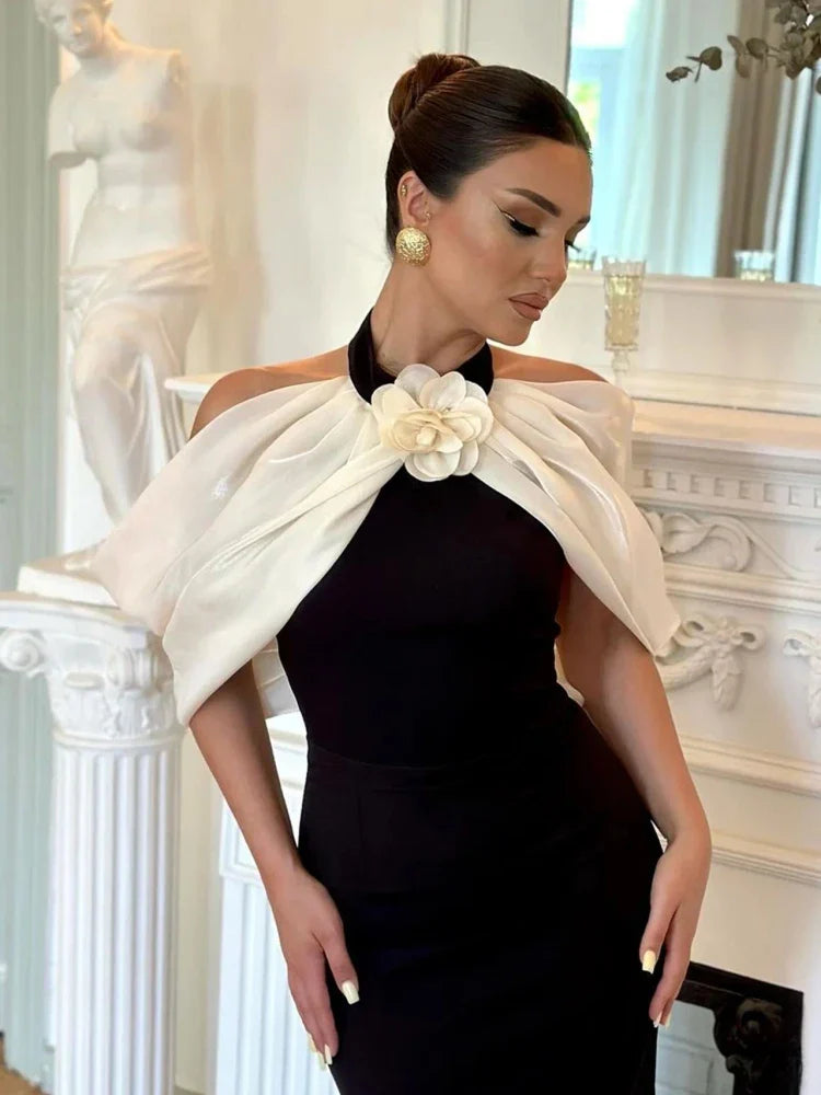 A woman standing in a classic pose with one hand on her hip, wearing an off-the-shoulder black dress with a 3D flower design, paired with white platform heels and a white belt.
