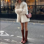 Front view of a woman in a cream faux fur coat with a notch lapel and long sleeves, standing outdoors.