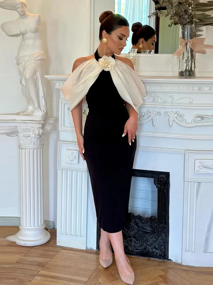 A woman standing in a classic pose with one hand on her hip, wearing an off-the-shoulder black dress with a 3D flower design, paired with white platform heels and a white belt.