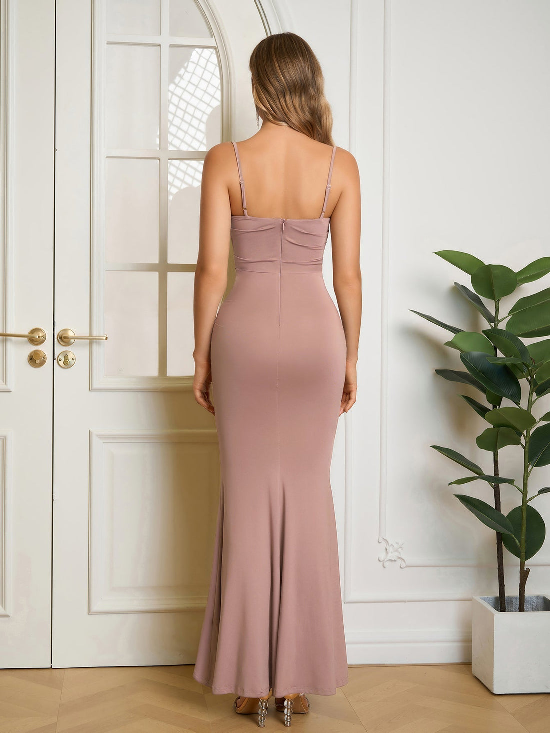 Woman wearing a long, taupe, spaghetti strap, ruched mermaid dress with a cowl neckline, standing with hand on hip.