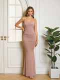 Woman wearing a long, taupe, spaghetti strap, ruched mermaid dress with a cowl neckline, standing with hand on hip.