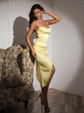 Woman in a yellow satin bodycon dress with a cowl neckline and spaghetti straps, standing next to a floral armchair.