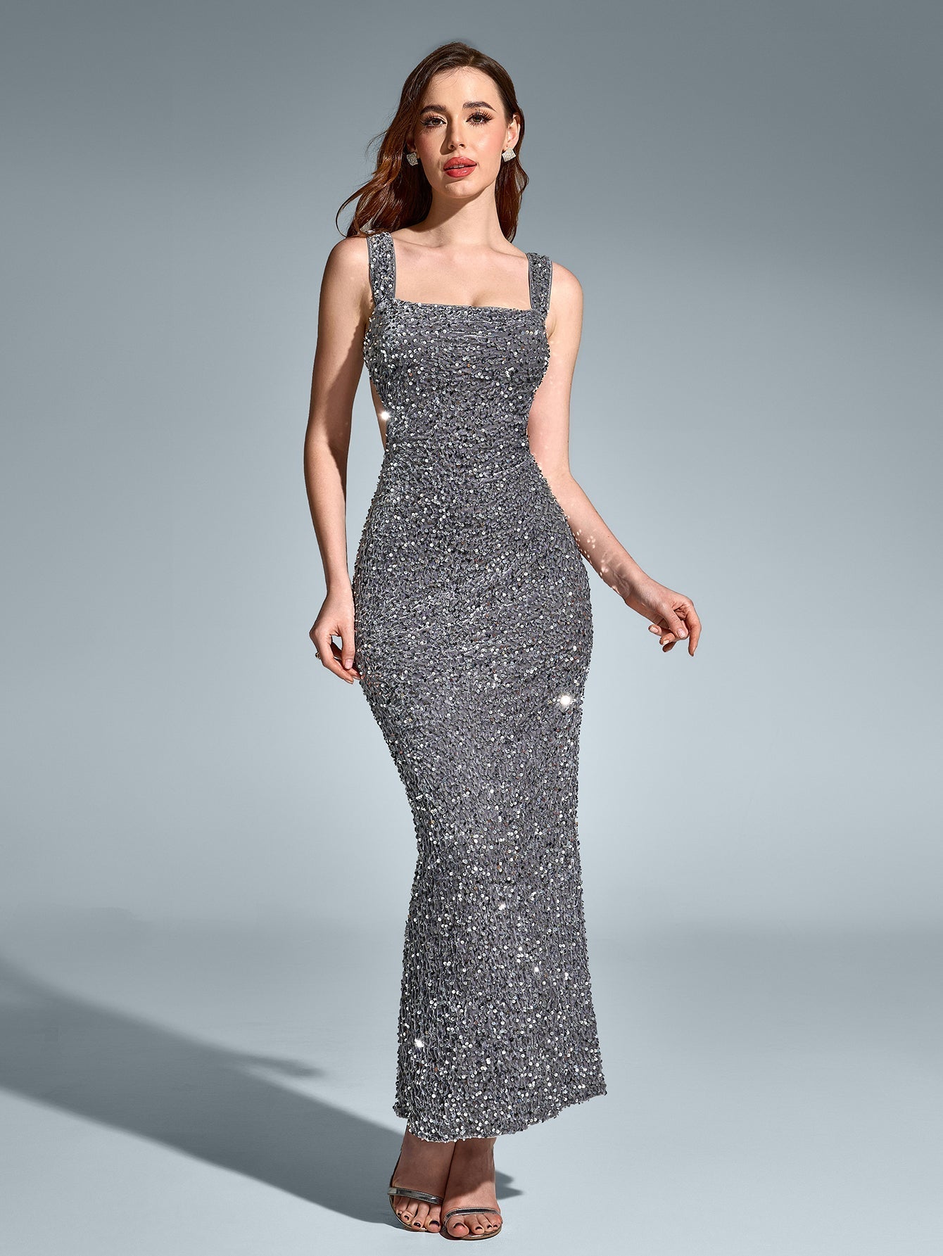 Full-length view of a woman wearing a sparkling silver sequined column gown with a square neckline and sleeveless straps, against a grey background.
