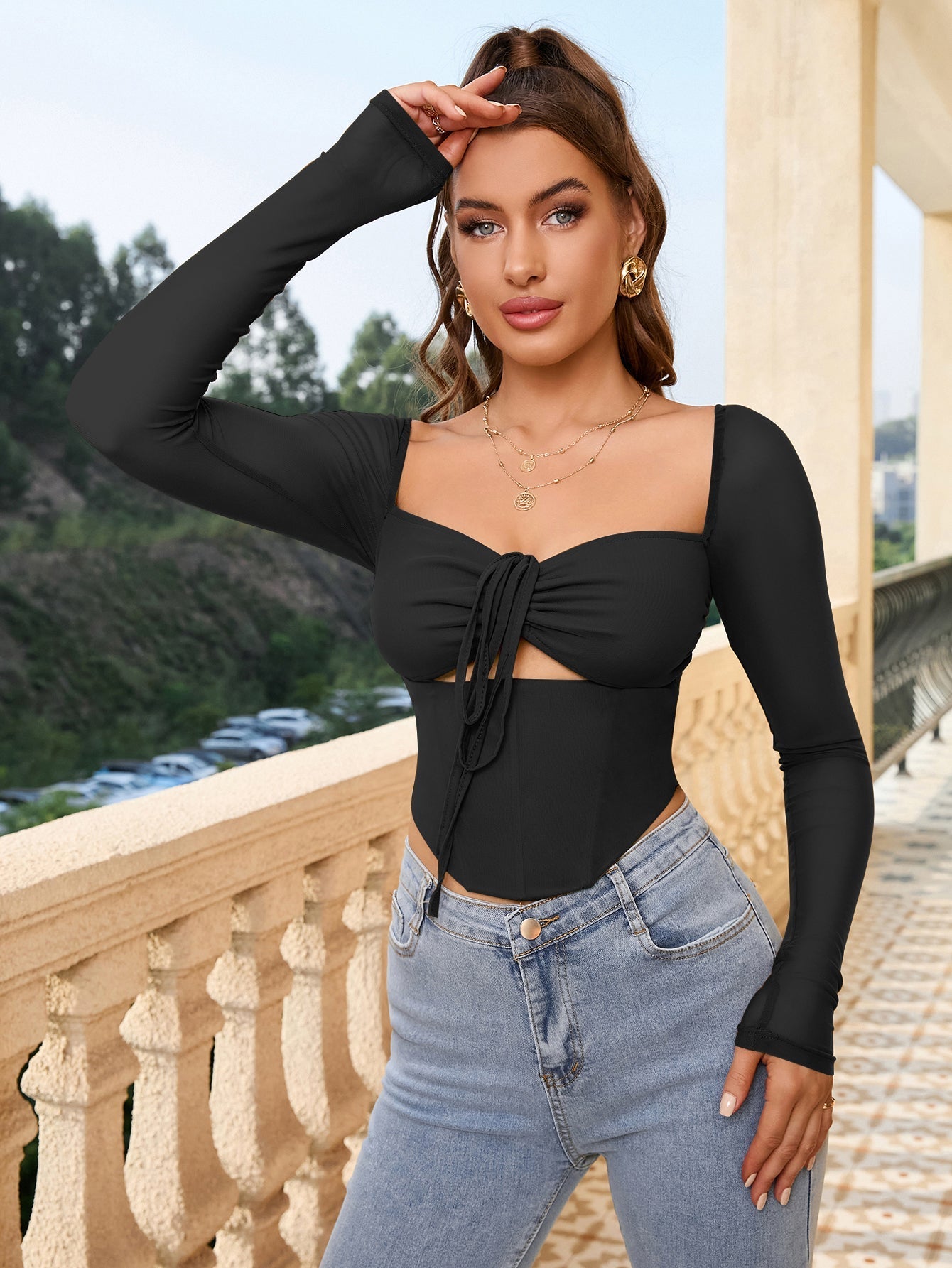 Woman in a black long-sleeve cropped top with a square neckline and tie detail, touching her hair.