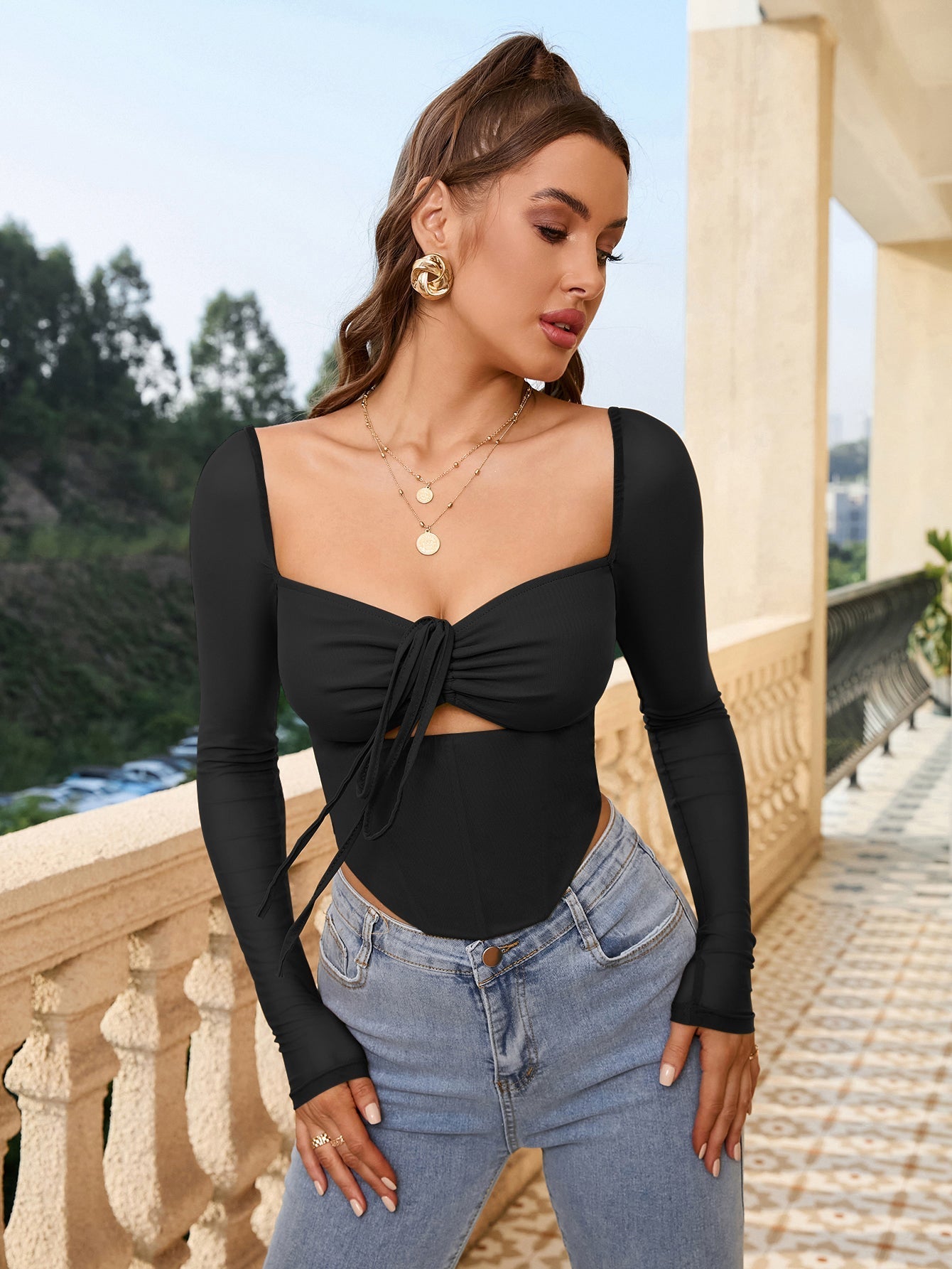 Woman in a black long-sleeve cropped top with a square neckline and tie detail, posing outdoors.