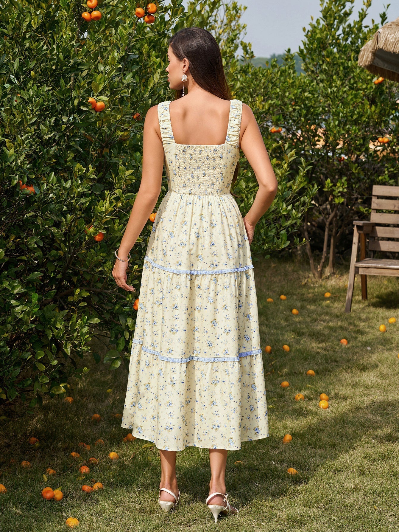 Woman in a straw hat wearing a yellow floral print midi dress and holding a basket of fruit, standing in an orchard.