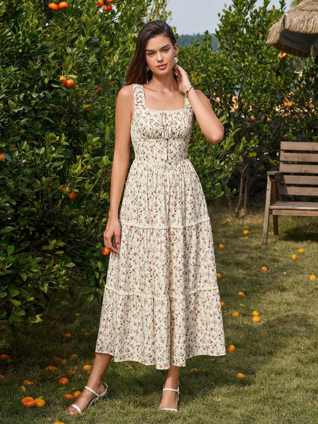 Woman wearing a yellow floral print sleeveless A-line midi dress with a square neckline and smocked bodice in an outdoor setting with citrus trees.