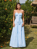 Woman wearing a light blue floral print strapless maxi dress with a bow detail at the neckline, standing in an orange grove.