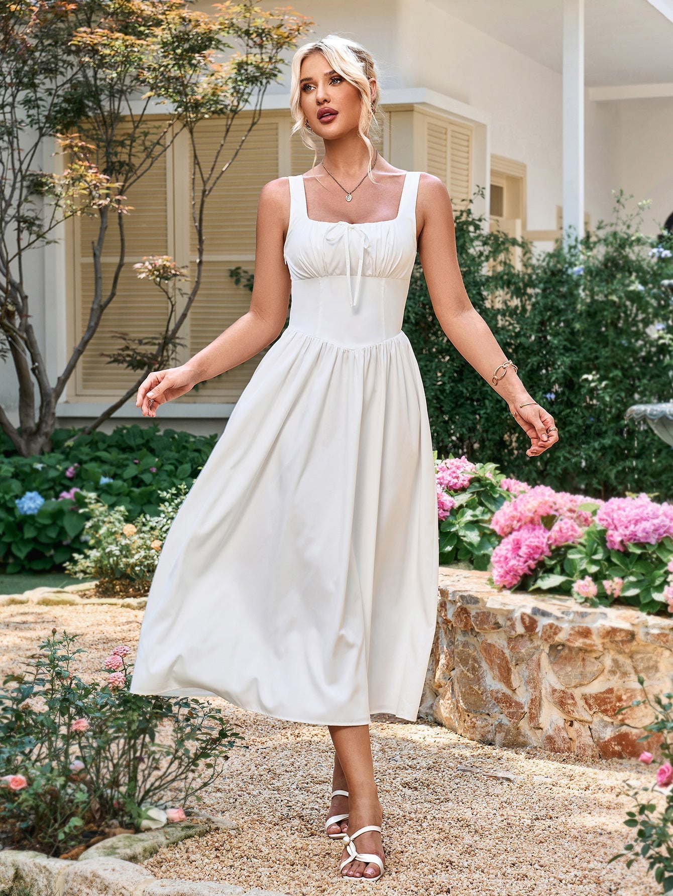 woman wearing a white sleeveless midi dress with a square neckline and tie detail, standing in a garden