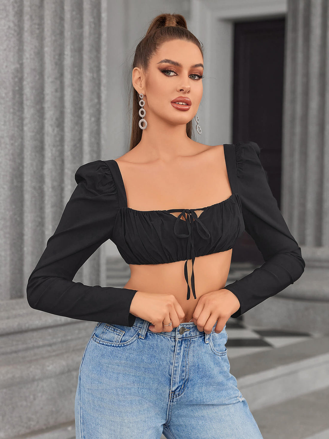 Woman wearing a black cropped long-sleeve top with a square neckline and tie detail, paired with blue jeans.