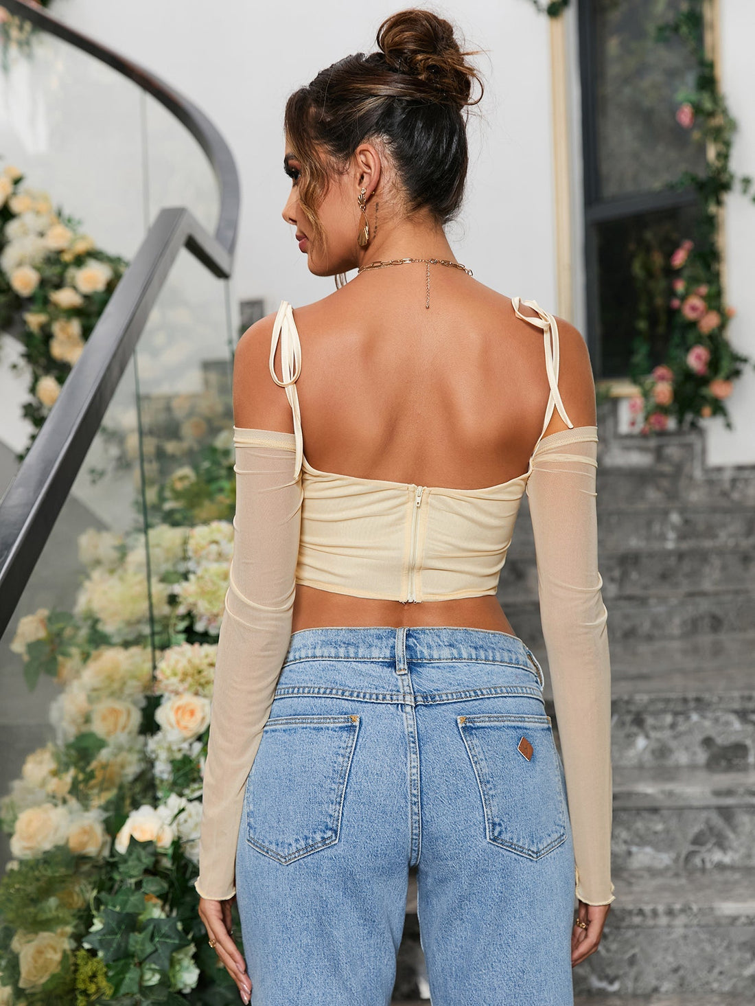 Woman wearing a light yellow ruched off-the-shoulder long-sleeve crop top with tie shoulder straps and a pointed hemline, paired with light wash denim jeans.
