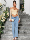 Full-body shot of a woman wearing a light yellow ruched off-the-shoulder long-sleeve crop top with tie shoulder straps and a pointed hemline, paired with light wash denim jeans and white heeled sandals.