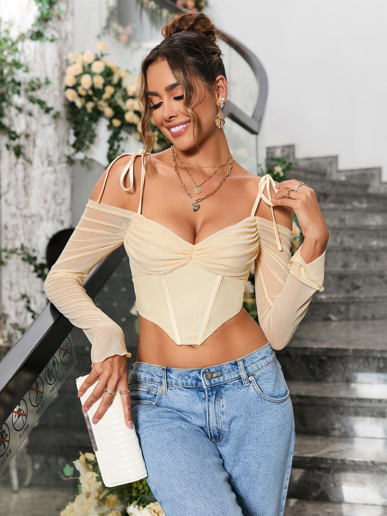 Close-up of a woman wearing a light yellow ruched off-the-shoulder long-sleeve crop top with tie shoulder straps and a pointed hemline, styled with layered necklaces.