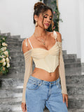 Woman wearing a light yellow ruched off-the-shoulder long-sleeve crop top with tie shoulder straps and a pointed hemline, showing off the ruched bodice detail, paired with light wash denim jeans.