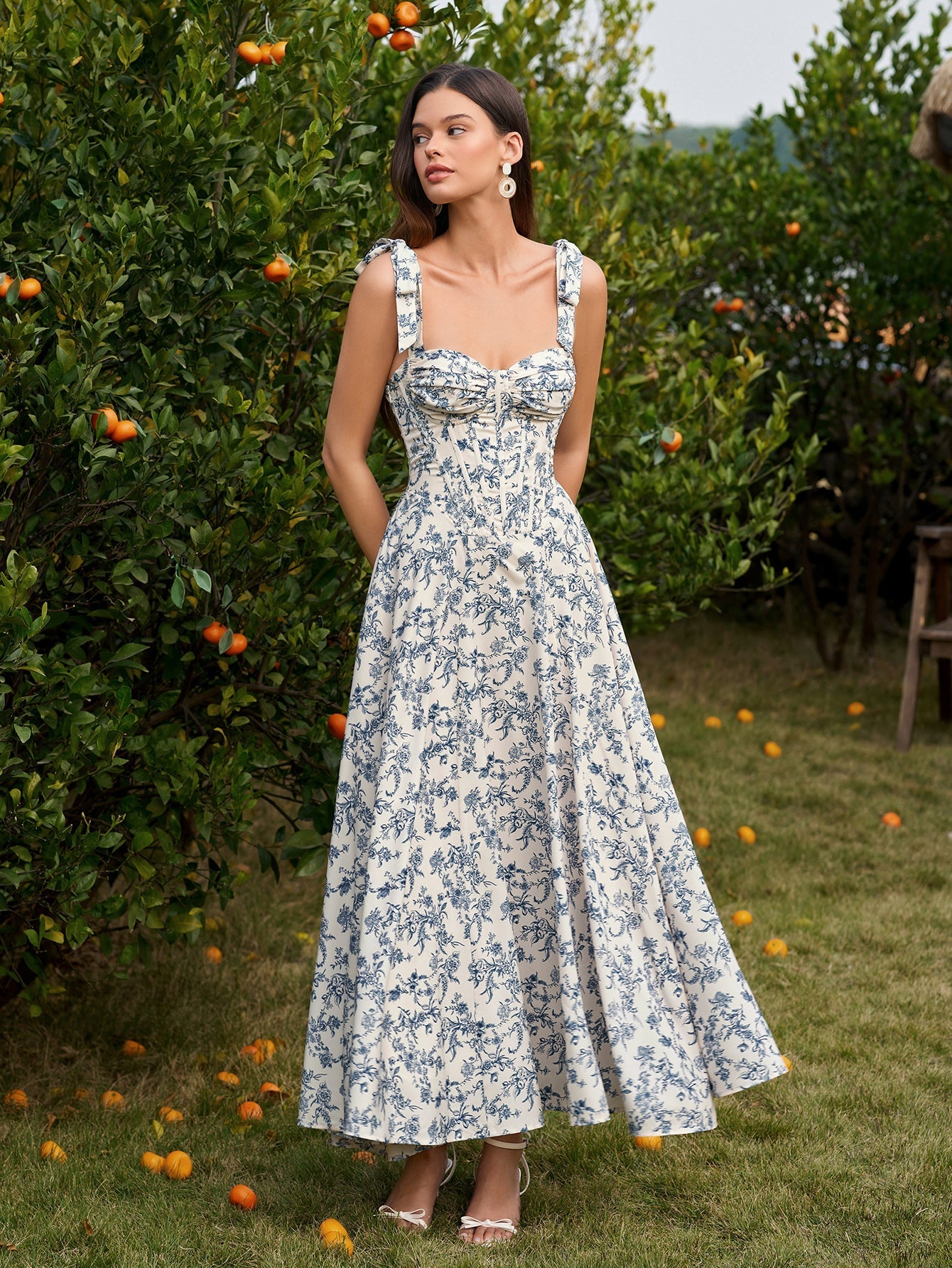 Full body shot of a woman in a floral print midi dress with a sweetheart neckline and tie shoulder straps, standing outdoors.
