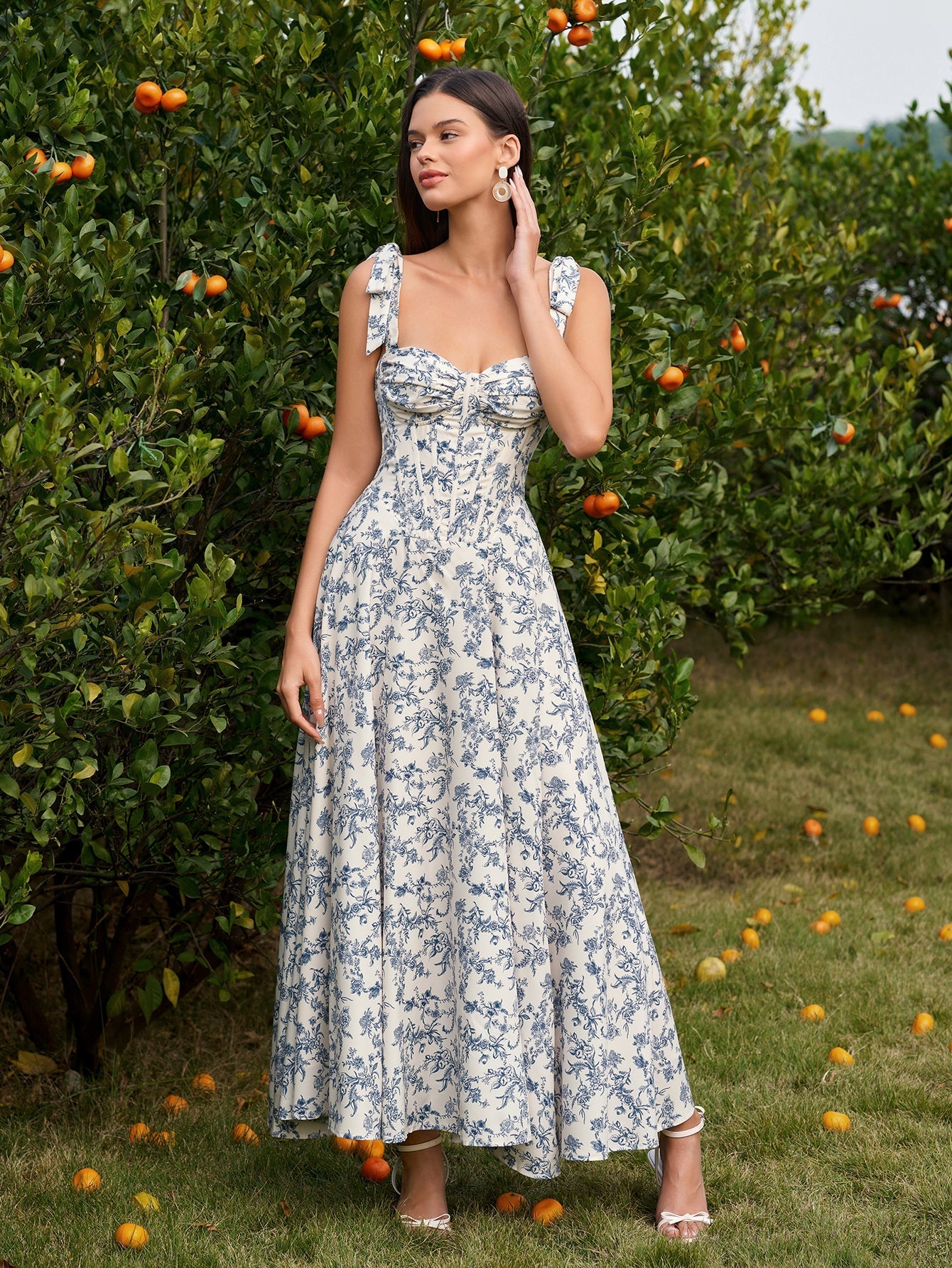 Woman wearing a blue and white floral print midi dress with a sweetheart neckline and tie shoulder straps, standing in front of an orange tree.