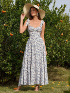 Woman wearing a straw hat and a floral print midi dress with a sweetheart neckline and tie shoulder straps, standing in front of an orange tree.