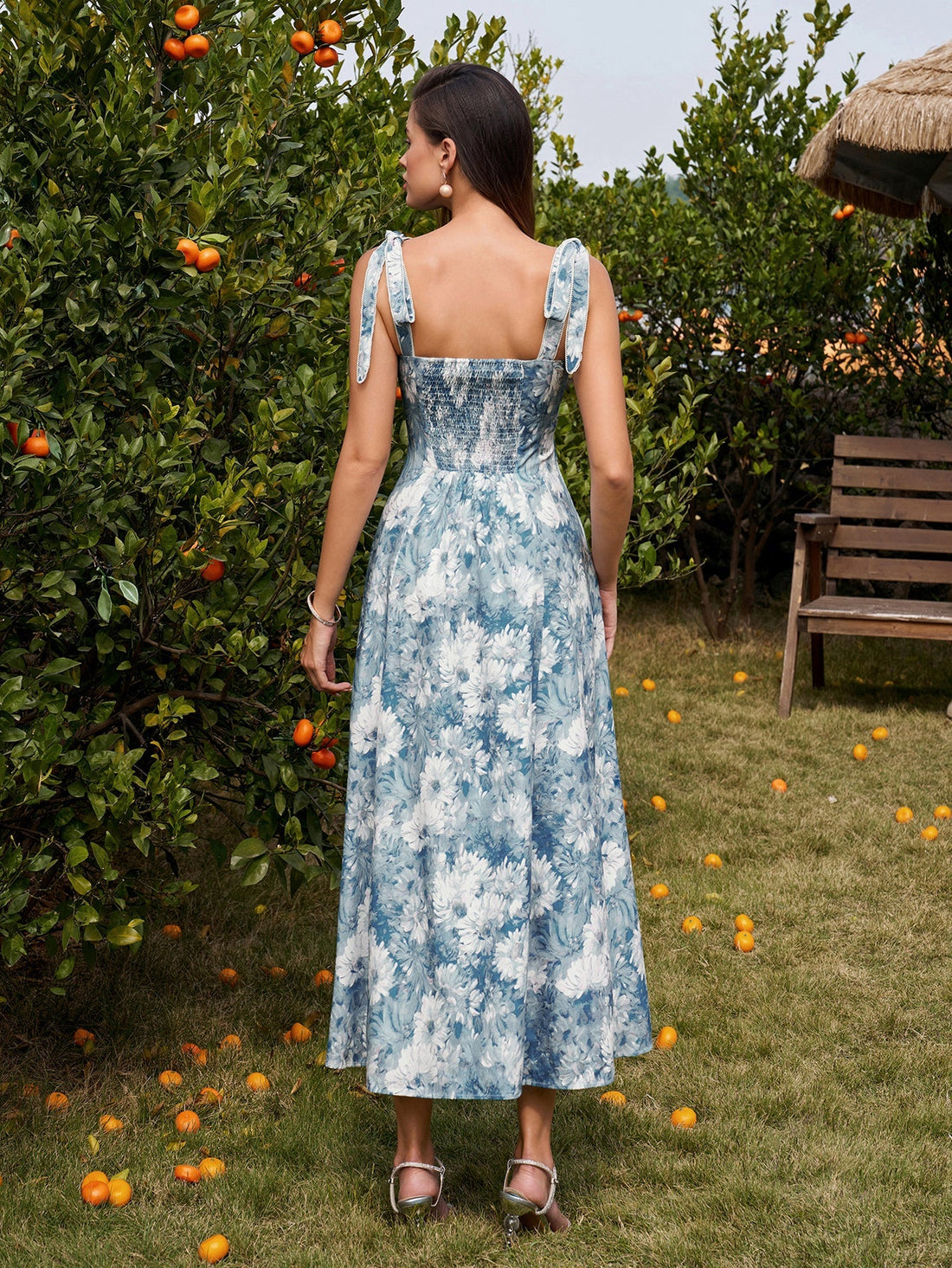Woman wearing a blue and white floral print midi dress with tie shoulder straps, standing in an orchard.