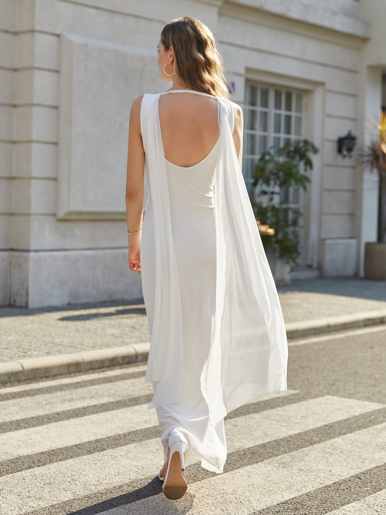 Back view of a woman in a long white sleeveless formal dress with an open back and flowing cape detail.