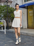 Woman walking in a white V-neck sleeveless crop top and white pleated mini skirt, outdoors.