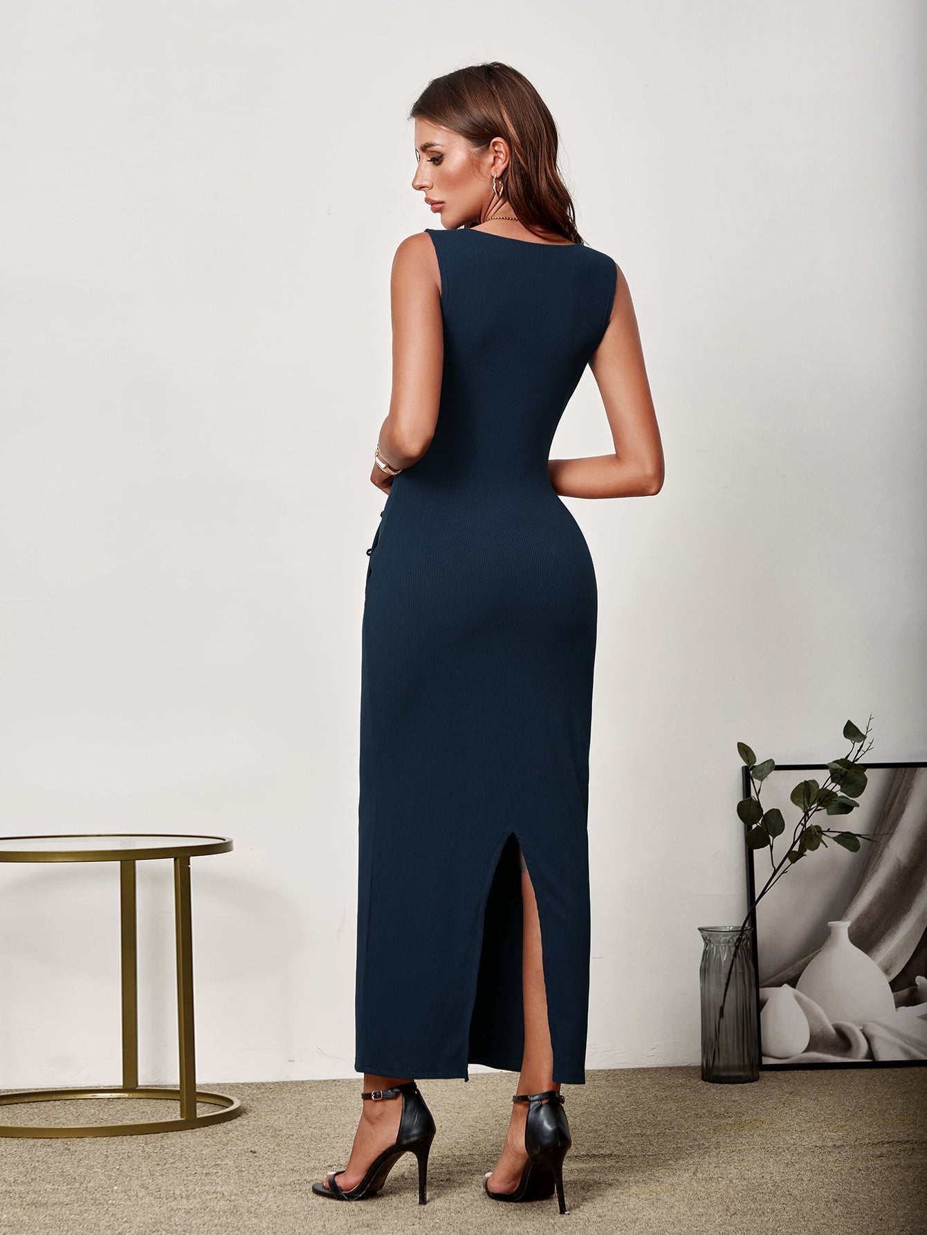 Back view of a woman in a navy blue sleeveless V-neck maxi sheath dress with button detailing and a side slit.