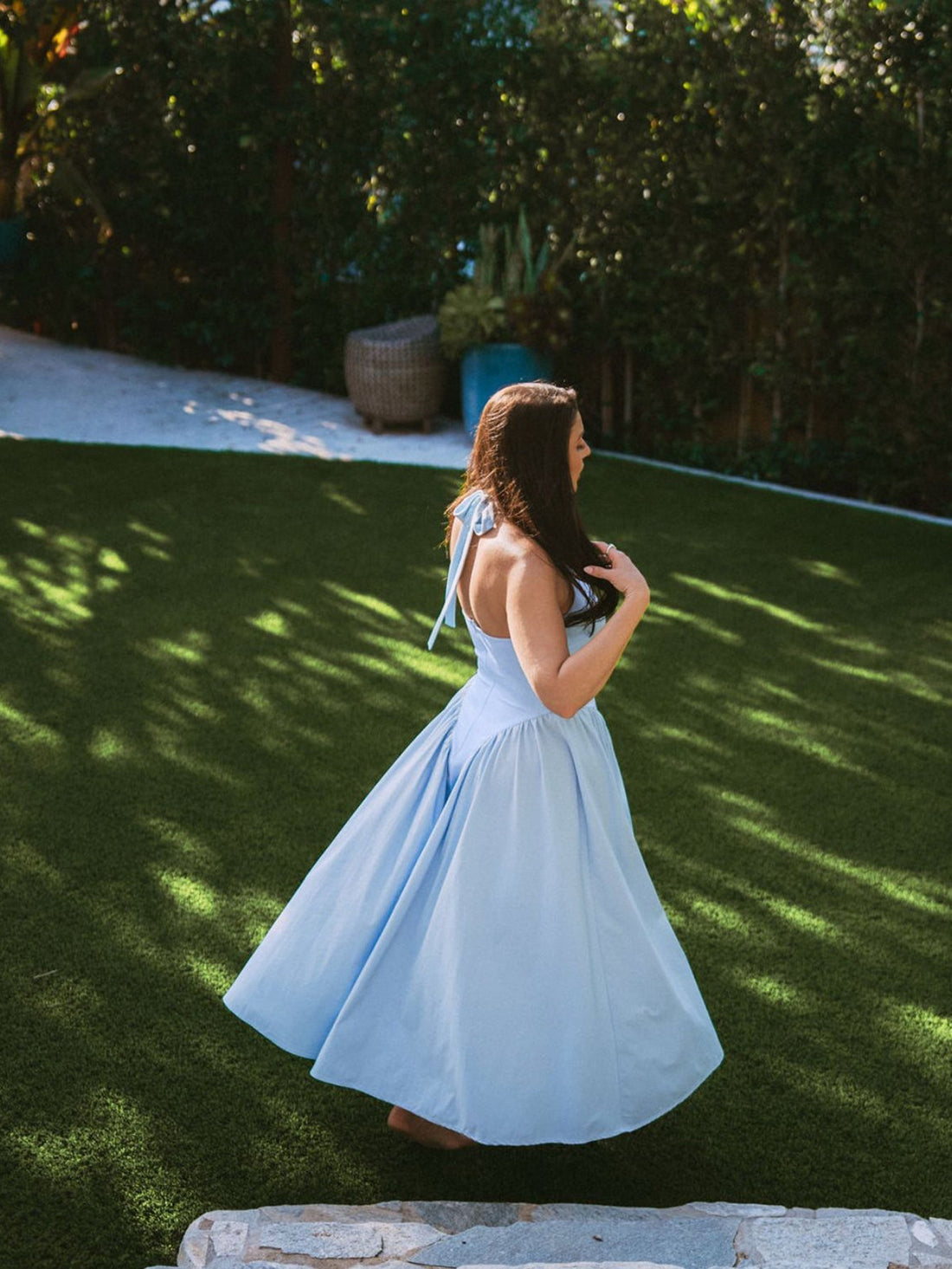 Woman in a light blue halter neck midi dress with a full skirt, standing indoors near a large window.