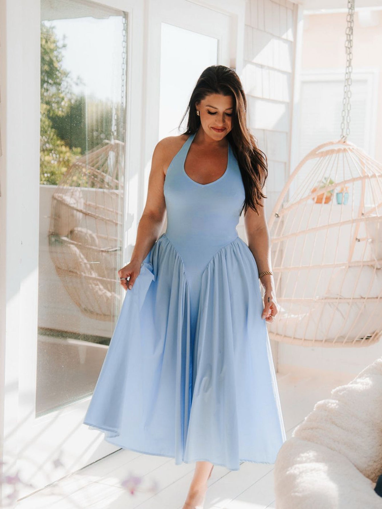 Woman in a light blue halter neck midi dress with a full skirt, standing indoors near a large window.