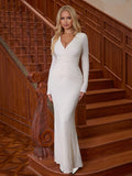 Woman wearing a white long-sleeved textured knit mermaid gown with a v-neckline and ruching detail, standing on a wooden staircase.