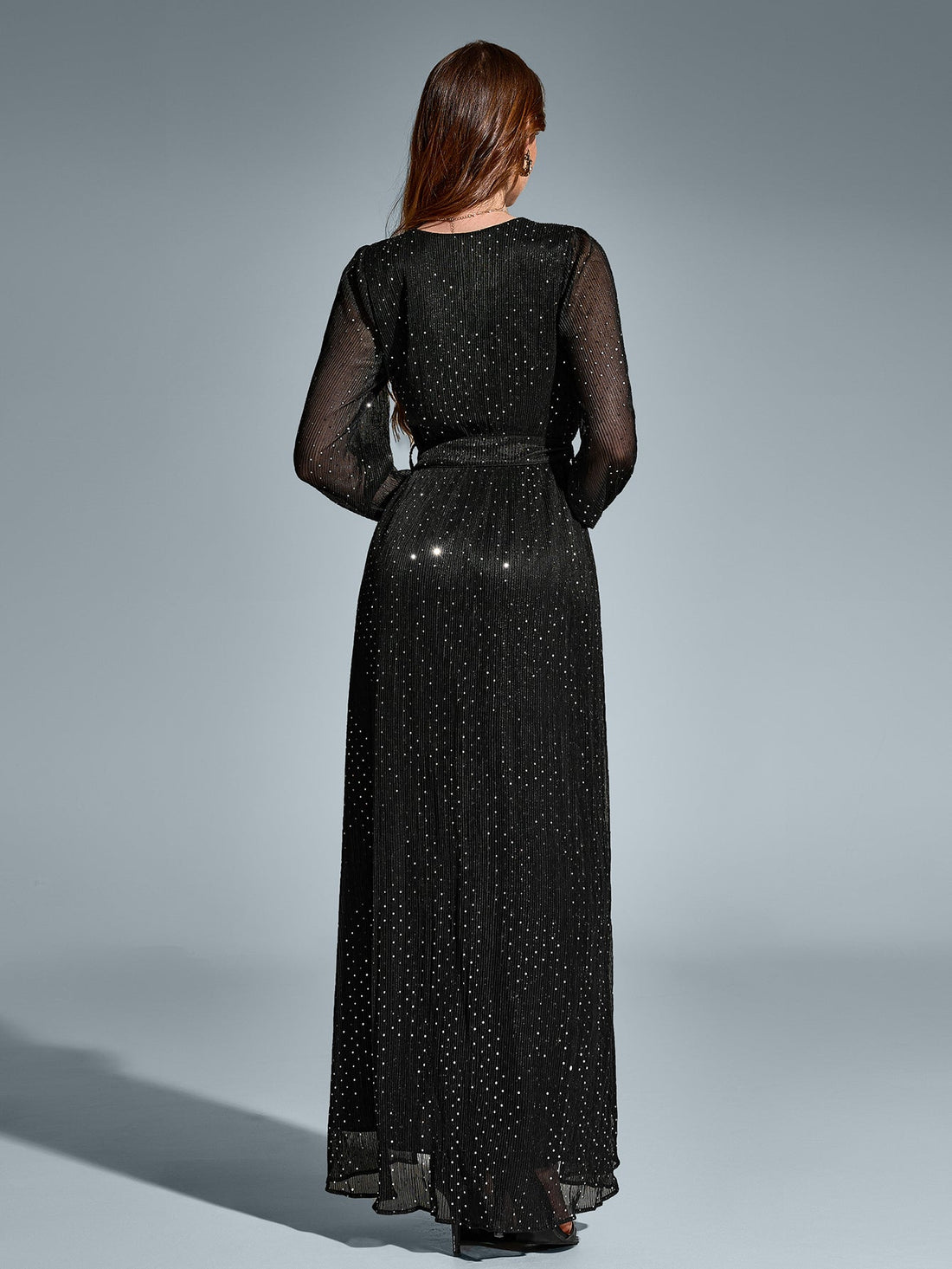 Woman in a black long-sleeved maxi dress with a v-neck and high slit, adorned with scattered metallic dots, posing with one hand on hip.